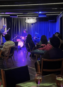 A band performs on stage in a dimly lit Bogotá bar as patrons seated at tables watch and listen.